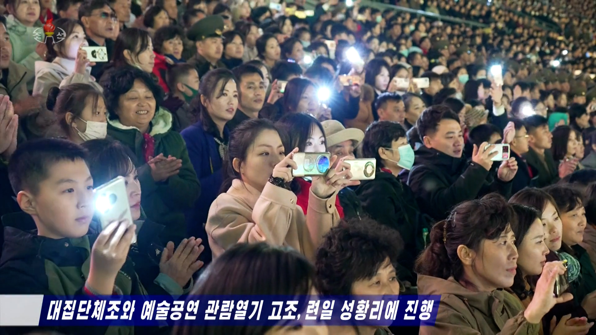 A large crowd of people in a stadium holding their smartphones to film an event, and use their smartphone flashlights